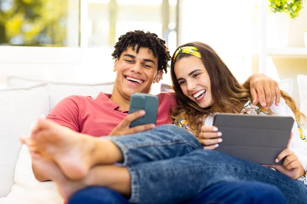Szczęśliwa Zróżnicowana Para Korzystająca Smartfona Tabletu Relaksująca Się Sofie Salonie — Zdjęcie stockowe