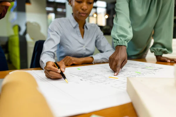 Midsection of african american female colleagues analyzing blueprint on desk in office. Unaltered, architecture, construction industry, design, teamwork, planning, gesture, hand, pen and business.