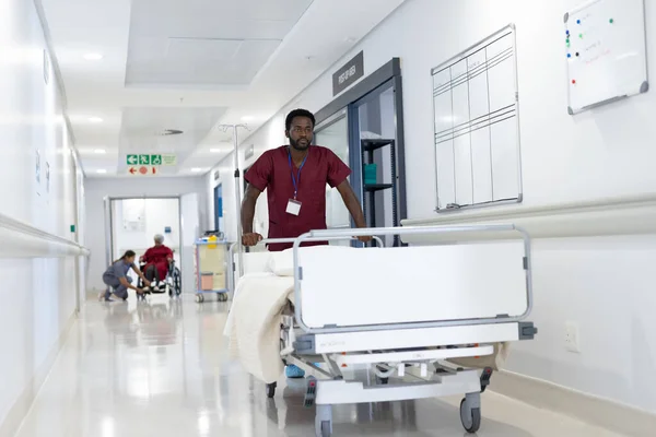 アフリカ系アメリカ人男性医療従事者が病院の廊下で空のベッドを押している 医療サービス — ストック写真
