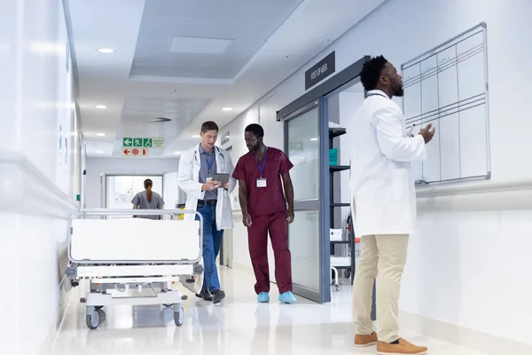Dwóch Różnych Lekarzy Używa Tabletów Rozmawia Zatłoczonym Szpitalnym Korytarzu Usługi — Zdjęcie stockowe