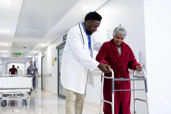 Diverse male doctor helping senior female patient use walking frame in hospital corridor, copy space. Hospital, medical and healthcare services.