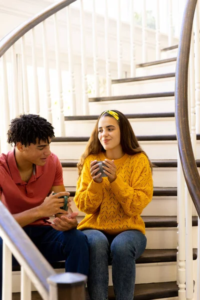 Dwurasowa Młoda Para Trzymająca Kubki Kawy Rozmawiająca Siedząc Schodach Domu — Zdjęcie stockowe
