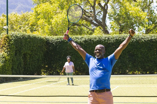 stock image Happy senior african american couple with tennis rackets, man celebrating on sunny tennis court. Senior lifestyle, retirement, sport, summer, fitness, hobbies and leisure activities.