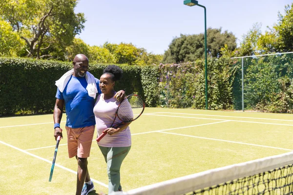 日当たりの良い芝生のコート コピースペースを歩いてテニスラケット付きの幸せなシニアアフリカ系アメリカ人のカップル シニアライフスタイル スポーツ フィットネス 趣味やレジャー活動 — ストック写真