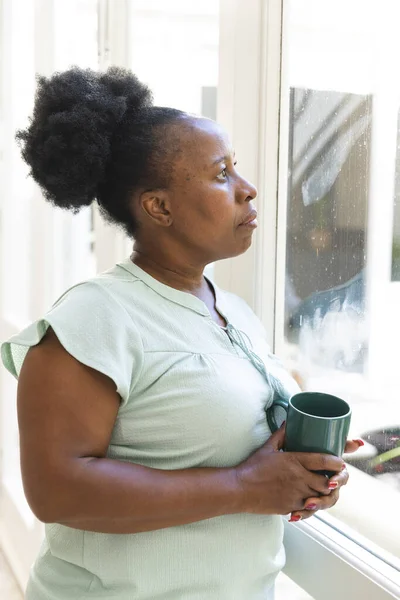 Przemyślana Starsza Afroamerykanka Pijąca Kawę Wyglądająca Przez Okno Domu Seniorski — Zdjęcie stockowe