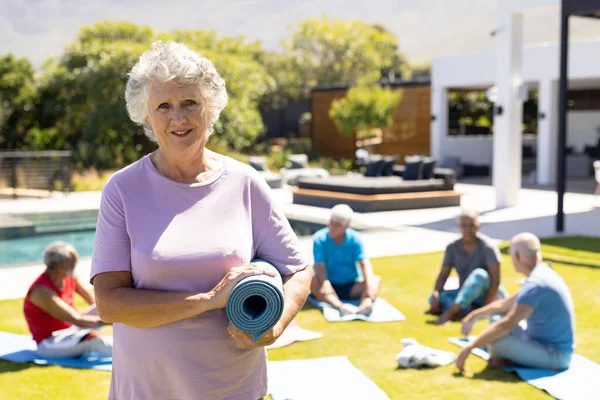 Portrait of happy senior caucasian woman holding yoga mat, with diverse yoga group in sunny garden. Senior lifestyle, active retirement, friendship, health and wellbeing, unaltered.