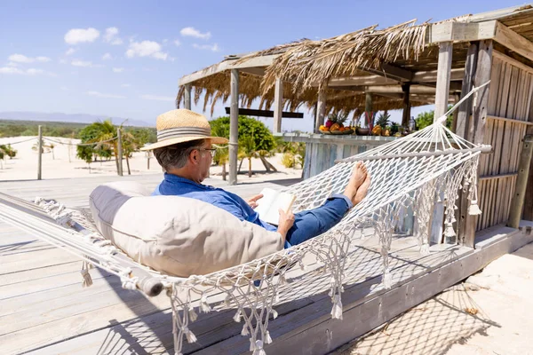 帽子をかぶって読書をしながら浜のハンモックに横たわっている白人の先輩 変更されていない 観光リゾート リラクゼーション 楽しみ 夏と趣味 — ストック写真