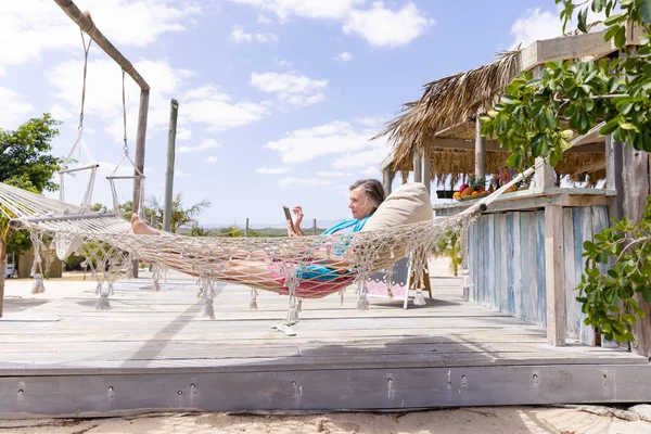 デジタルタブレットを使用し 空に対してビーチでハンモックに休んで白人のシニア女性の側のビュー 変更されていない 観光リゾート リラクゼーション 楽しみ 夏と技術 — ストック写真