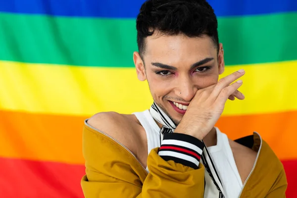 Portrait of happy biracial transgender man looking at camera with rainbow flag in background. Gender, lgbtq, pride and lifestyle, unaltered.