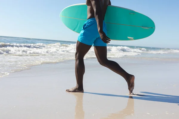適切なアフリカ系アメリカ人の男性の海に日当たりの良いビーチを歩いてサーフボードを運ぶの低セクション 健康的なライフスタイル スポーツ サーフィンや休暇 変更なし — ストック写真