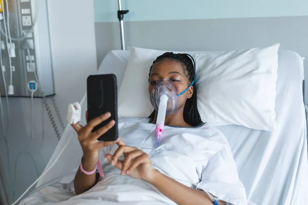 アフリカ系アメリカ人女性酸素マスク患者で ベッドに横になり 病院の部屋でスマートフォンを使用しています コミュニケーション 医学と医療 変更なし — ストック写真