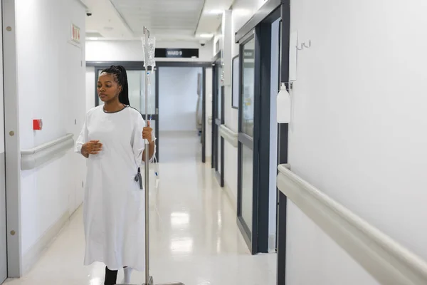 非洲裔美国女性患者身穿医院礼服 手握滴水 走在走廊上 医药和保健 未加改动 — 图库照片