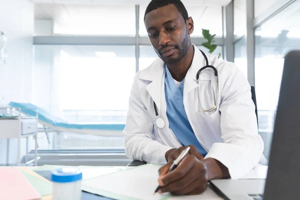 アフリカ系アメリカ人の男性医師は 研究室のコートと聴診器を着て 机の上に座り メモをとる 医療と仕事 変更なし — ストック写真