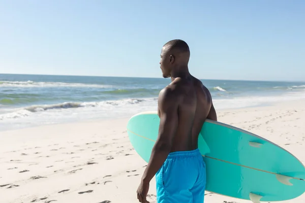日当たりの良いビーチでサーフボードを運ぶアフリカ系アメリカ人の男に合わせて 海を見てください 健康的なライフスタイル スポーツ サーフィンや休暇 変更なし — ストック写真