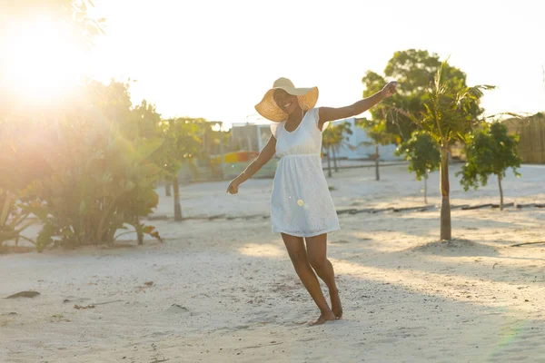 戴着太阳帽 身穿白色海滩礼服的快乐的非洲裔美国女人在阳光灿烂的海滩上跳舞 放松和假期 — 图库照片