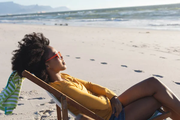 戴着太阳镜的快乐的非洲裔美国女人在海上阳光明媚的海滩上坐在甲板上休息 闲暇时间和假期 — 图库照片
