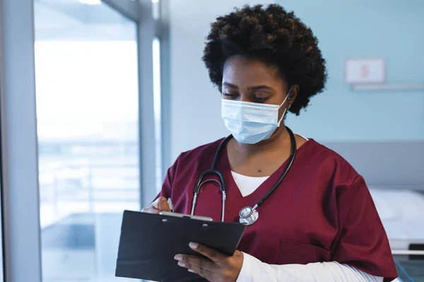 African american female doctor wearing scrubs and face mask, holding clipboard and writing. Hospital, medicine, healthcare and work, unaltered.