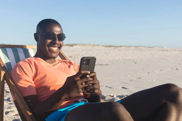 Szczęśliwy Amerykanin Okularach Przeciwsłonecznych Siedzący Leżaku Korzystający Smartfona Słonecznej Plaży — Zdjęcie stockowe