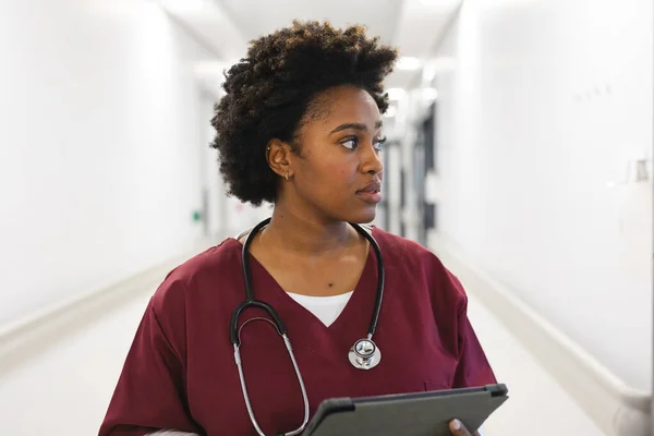 アフリカ系アメリカ人の女性医師は 病院の廊下にタブレットを使用して スクラブと聴診器を身に着けています コミュニケーション 医療と仕事 変更なし — ストック写真