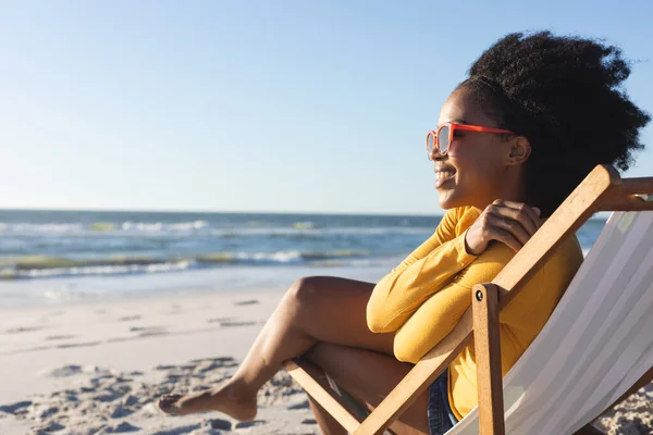 戴着太阳镜的快乐的非洲裔美国女人坐在甲板上 微笑着坐在海上阳光灿烂的海滩上 闲暇时间和假期 — 图库照片