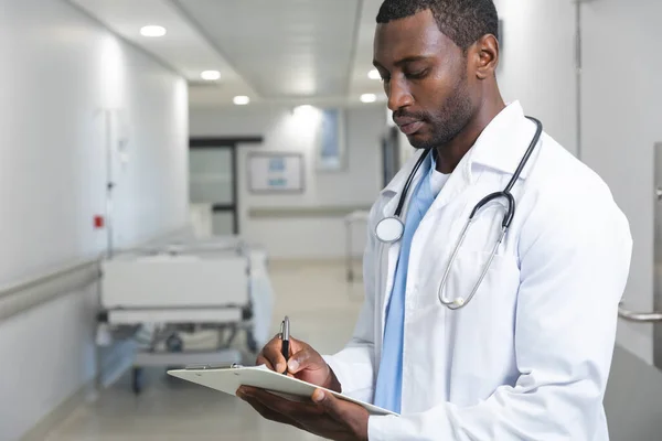 Médico Americano Africano Vestindo Jaleco Estetoscópio Segurando Prancheta Escrevendo Hospital — Fotografia de Stock