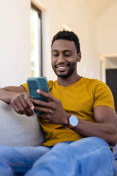 幸せなアフリカ系アメリカ人男性がリビングルームでスマートフォンを使用しています リラクゼーション ライフスタイル コミュニケーション 国内生活 変わらない — ストック写真