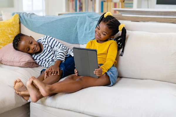 非洲裔美国人快乐的姐姐坐在客厅的沙发上 一边用数码平板电脑 一边和哥哥坐在一起 不变的家庭 无线技术和家庭观念 — 图库照片
