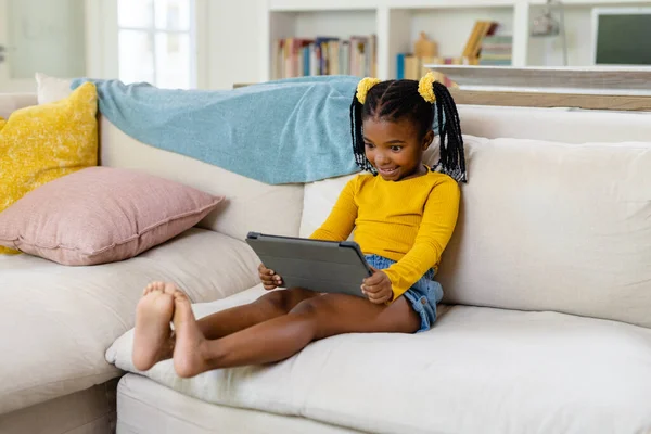 リビングルームでソファに座っている間 アフリカ系アメリカ人の笑顔の女の子はデジタルタブレットを使用しています 変更されていない 子供時代 ライフスタイル 無線技術 リラクゼーション 幸せと家庭の概念 — ストック写真