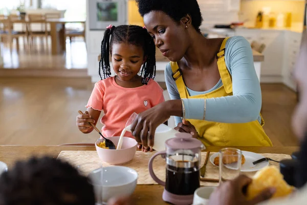 アフリカ系アメリカ人の母親は 自宅のダイニングテーブルで娘のための朝食のシリアルに牛乳を注ぐ 変更されていない ライフスタイル 一緒に 子供時代 食べ物や家庭の概念 — ストック写真