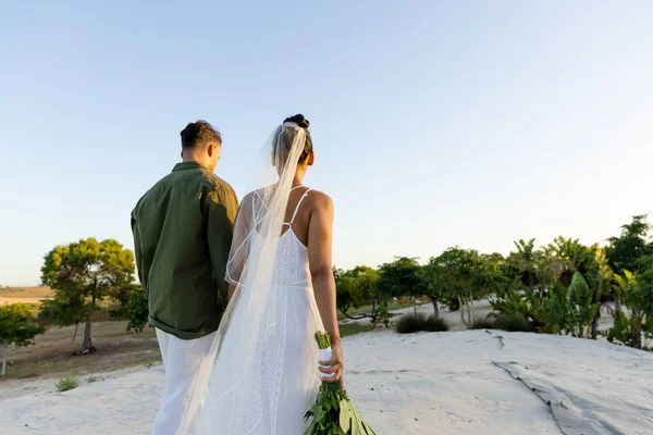 澄んだ空に向かって木々の間の砂浜を歩いている新婚夫婦の後ろの景色 コピースペース 変更されていない 一緒に 宛先結婚式 イベント お祝い — ストック写真