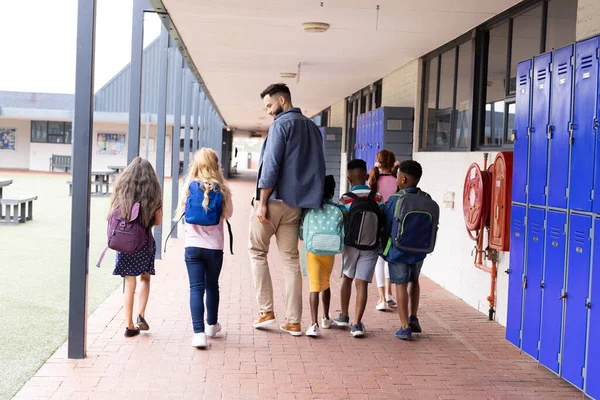 小学校の廊下を歩く多様な男性教師と子供たちのリアビュー コピースペース コミュニティ 小学校 学習コンセプト — ストック写真