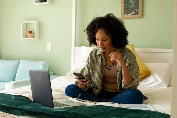 Mujer Biracial Haciendo Papeleo Usando Portátil Teléfono Inteligente Cama Dormitorio — Foto de Stock