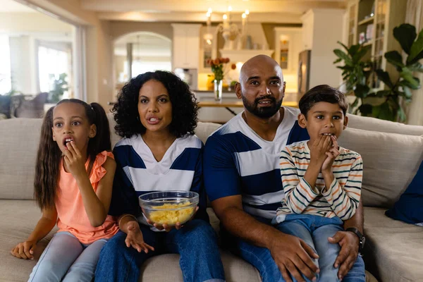 Focussed biracial family sitting on couch eating snacks and watching tv. Family, sport, entertainment, free time, togetherness, lifestyle and domestic life, unaltered.