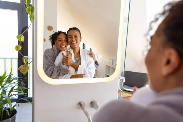 Feliz Casal Lésbico Biracial Roupões Banho Abraçando Olhando Espelho Banheiro — Fotografia de Stock