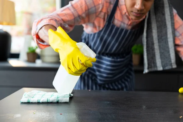 Midsection of biracial man wearing apron and rubber gloves cleaning countertop in sunny kitchen. Cleaning, hygiene, healthy lifestyle and domestic life, unaltered.