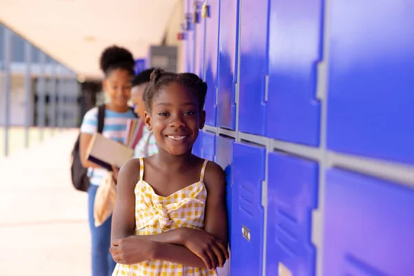 Portrait of happy african american schoolgirl standing next to locker at school. Education, childhood, elementary school and learning concept.