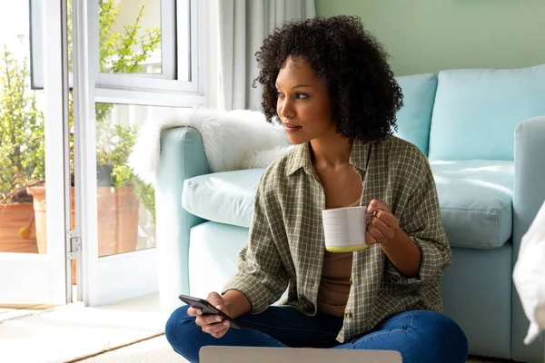 Donna Biracial Utilizzando Smartphone Tenendo Tazza Caffè Seduto Sul Pavimento — Foto Stock