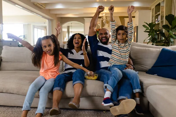 Happy biracial family sitting on couch with snacks, watching tv and cheering. Family, sport, entertainment, free time, togetherness, lifestyle and domestic life, unaltered.