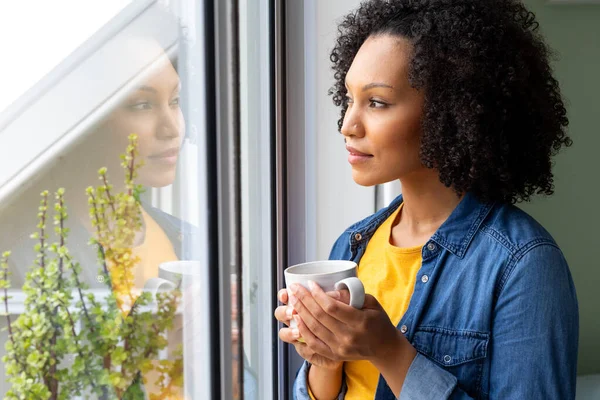 Dwurasowa Kobieta Trzymająca Filiżankę Kawy Wyglądająca Przez Okno Domu Relaks — Zdjęcie stockowe