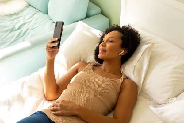 スマートフォンを使ってベッドの上に寝そべっているイヤホン付きの幸せな出産女性 リラクゼーション ライフスタイル コミュニケーション 国内生活 変わらない — ストック写真
