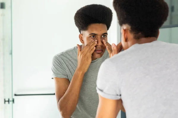 Homem Biracial Olhando Espelho Inspecionando Rosto Olhos Banheiro Ensolarado Autocuidado — Fotografia de Stock