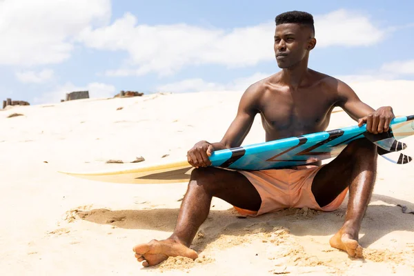 Focused Biracial Man Holding Surfboard Sitting Sunny Beach Looking Away — Stock Photo, Image