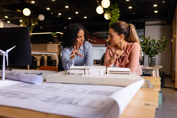 Diverse female architects in discussion using model and blueprints in casual office meeting. Casual office, teamwork, business, lifestyle, creative work, design and work, unaltered.
