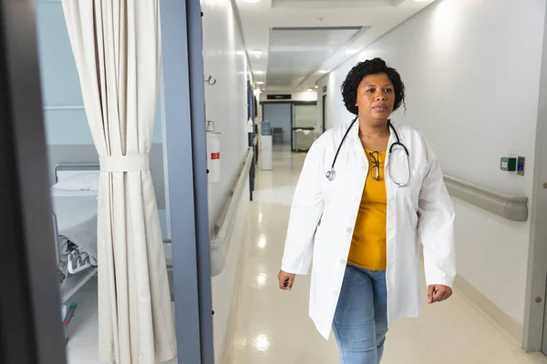 アフリカ系アメリカ人女性医師は 病院の廊下を歩くラボコートとステスコープを身に着けています 変わらない — ストック写真