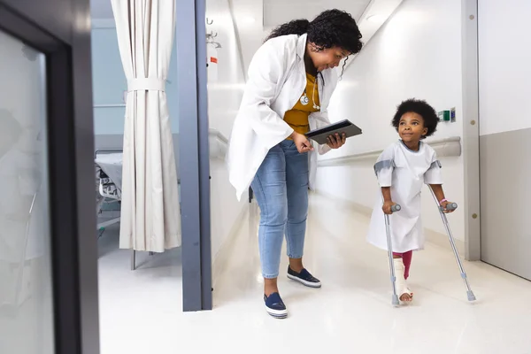 非洲裔美国女医生和女病人抱着拐杖在医院走廊里行走 医药和保健 未加改动 — 图库照片