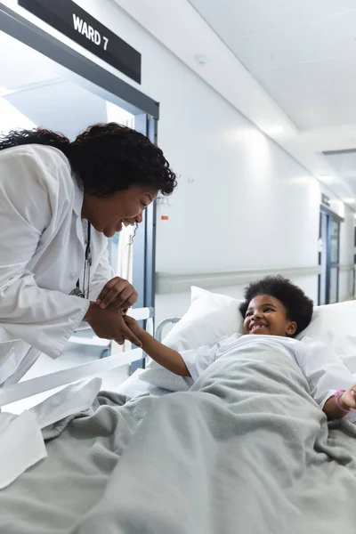 アフリカ系アメリカ人女性医師が病院の廊下でベッドに横たわっている少女患者と話している 変わらない — ストック写真