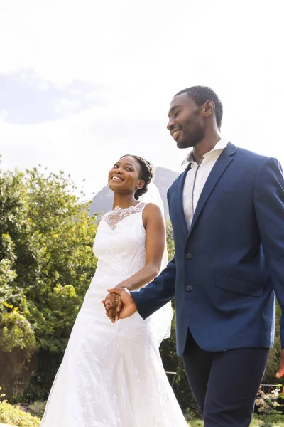 Smiling african american bride and groom holding hands, walking in sunny garden, copy space. Marriage, romance, summer, tradition, ceremony and lifestyle, unaltered.