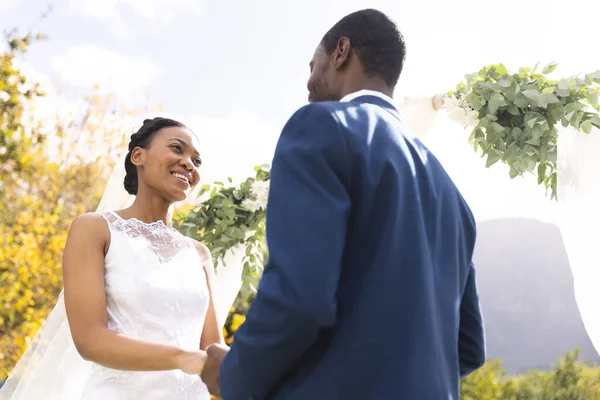 Happy african american bride and groom holding hands at wedding ceremony in sunny garden. Marriage, romance, summer, tradition, ceremony and lifestyle, unaltered.