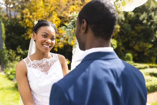 Happy african american bride and groom smiling at wedding ceremony in sunny garden. Marriage, romance, summer, tradition, ceremony and lifestyle, unaltered.