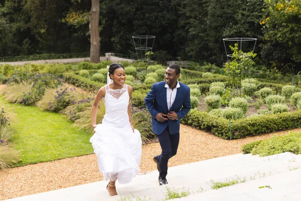 Happy african american bride and groom on wedding day running up steps together in sunny garden. Marriage, romance, summer, tradition, ceremony and lifestyle, unaltered.